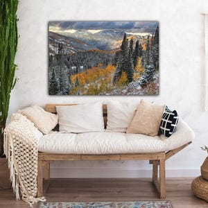 a cozy living room with a large, vibrant painting of a mountainous landscape hanging on the wall above a wooden bench. The painting showcases a breathtaking view of a forested mountain range with a mix of green and orange trees, creating a serene and picturesque scene.