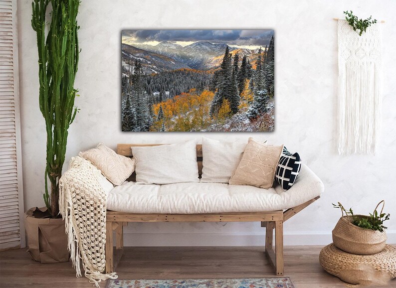 a cozy living room with a large, vibrant painting of a mountainous landscape hanging on the wall above a wooden bench. The painting showcases a breathtaking view of a forested mountain range with a mix of green and orange trees, creating a serene and picturesque scene.