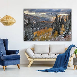 a cozy living room with a large landscape painting hanging on the wall, featuring a snowy mountain range with trees and fall foliage. The room is furnished with a blue armchair, a gray sofa, and a wooden side table with a potted plant. The overall atmosphere of the room is warm and inviting, with the natural elements of the painting complementing the comfortable furnishings.