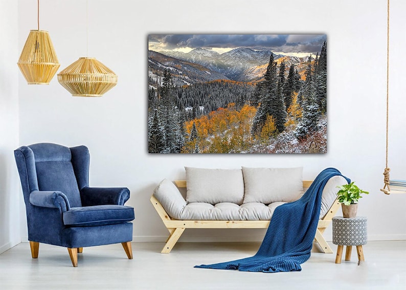 a cozy living room with a large landscape painting hanging on the wall, featuring a snowy mountain range with trees and fall foliage. The room is furnished with a blue armchair, a gray sofa, and a wooden side table with a potted plant. The overall atmosphere of the room is warm and inviting, with the natural elements of the painting complementing the comfortable furnishings.