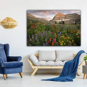 a serene living room with a large, vibrant painting of a mountain landscape hanging on the wall. The room is furnished with a blue armchair, a gray sofa, and a wooden bench. The room is decorated with pendant lights, a woven lampshade, and a potted plant, creating a cozy and inviting atmosphere.