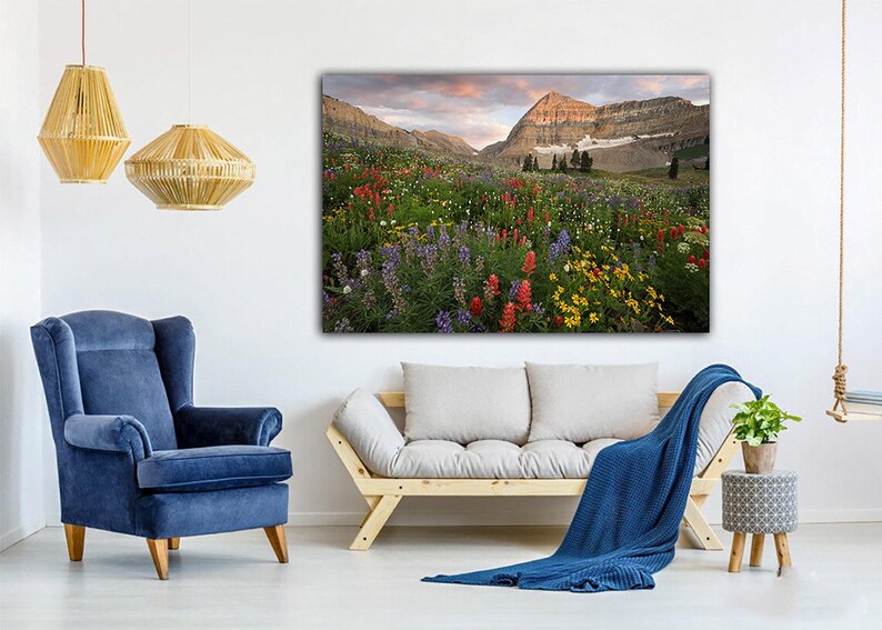 a serene living room with a large, vibrant painting of a mountain landscape hanging on the wall. The room is furnished with a blue armchair, a gray sofa, and a wooden bench. The room is decorated with pendant lights, a woven lampshade, and a potted plant, creating a cozy and inviting atmosphere.