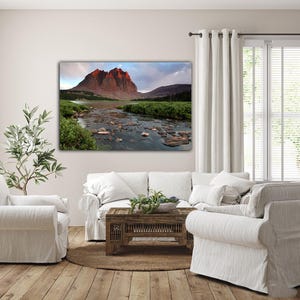 a serene living room with a large landscape painting of a mountain and river on the wall. The painting features a rocky mountain with a river flowing through it, surrounded by lush greenery. The room is furnished with a white sofa, a coffee table, and a potted plant, creating a cozy and inviting atmosphere.