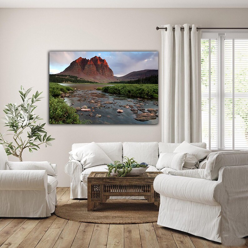 a serene living room with a large landscape painting of a mountain and river on the wall. The painting features a rocky mountain with a river flowing through it, surrounded by lush greenery. The room is furnished with a white sofa, a coffee table, and a potted plant, creating a cozy and inviting atmosphere.