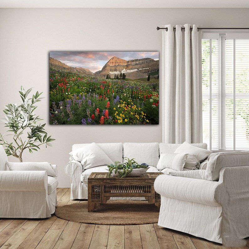 a serene living room with a large, vibrant painting of a mountain landscape hanging on the wall. The painting features a colorful array of flowers and trees, creating a picturesque scene. The room is furnished with a white sofa and chairs, and a wooden coffee table is placed in the center of the space. A large window allows natural light to flood the room, enhancing the overall ambiance.