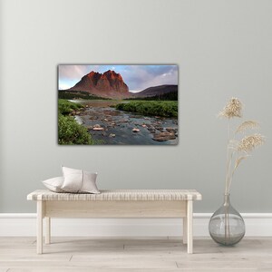 a large landscape painting hanging on a wall, depicting a mountain range with a river flowing through the valley. The painting is surrounded by a minimalist setting, with a bench and a vase of dried flowers placed on a wooden floor.