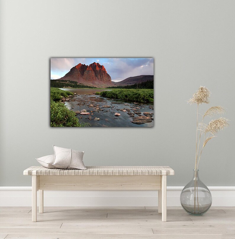 a large landscape painting hanging on a wall, depicting a mountain range with a river flowing through the valley. The painting is surrounded by a minimalist setting, with a bench and a vase of dried flowers placed on a wooden floor.