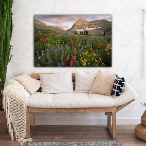 a cozy living room with a large, vibrant painting of a mountain landscape hanging on the wall above a wooden bench. The painting depicts a breathtaking view of a mountain range with colorful wildflowers in the foreground. The room is decorated with various plants and textiles, creating a warm and inviting atmosphere.