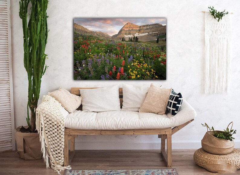 a cozy living room with a large, vibrant painting of a mountain landscape hanging on the wall above a wooden bench. The painting depicts a breathtaking view of a mountain range with colorful wildflowers in the foreground. The room is decorated with various plants and textiles, creating a warm and inviting atmosphere.