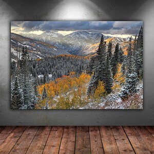 The image is a large framed landscape painting depicting a mountainous, forested area with a mix of snow-covered peaks and vibrant fall foliage.
