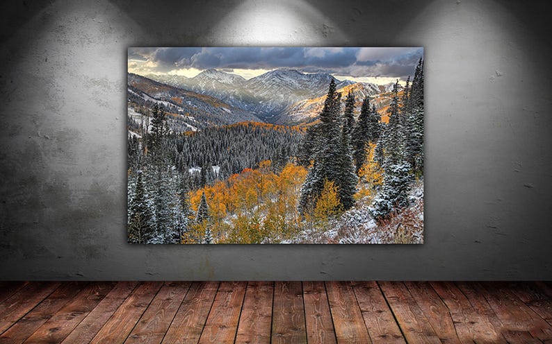 The image is a large framed landscape painting depicting a mountainous, forested area with a mix of snow-covered peaks and vibrant fall foliage.