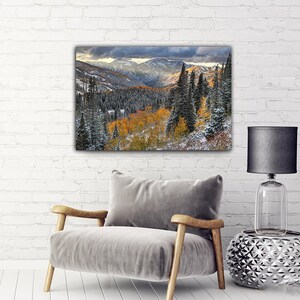 a cozy living room with a gray armchair, a silver side table, and a black lamp. Above the armchair, there is a large landscape painting depicting a mountainous, forested area with autumn foliage.