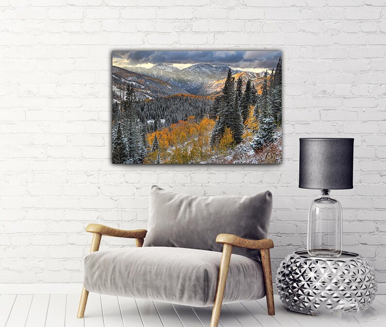 a cozy living room with a gray armchair, a silver side table, and a black lamp. Above the armchair, there is a large landscape painting depicting a mountainous, forested area with autumn foliage.