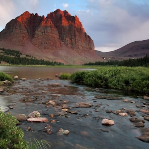 a serene natural landscape with a large, red rock formation rising majestically from a river, surrounded by lush greenery and a cloudy sky.