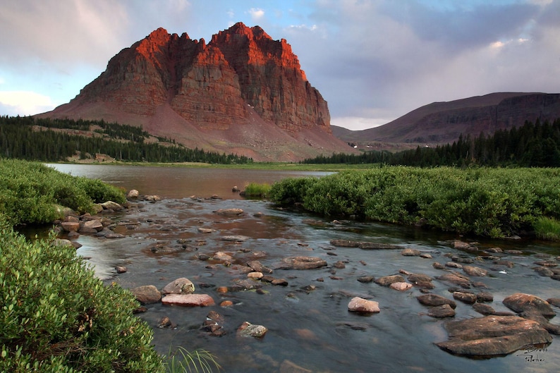 a serene natural landscape with a large, red rock formation rising majestically from a river, surrounded by lush greenery and a cloudy sky.