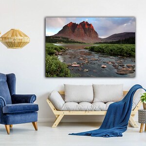 a living room with a large landscape painting of a mountain river and lush greenery hanging on the wall. The room is furnished with a blue armchair, a white sofa, and a wooden side table with a plant. The overall atmosphere of the room is calm and serene, with the natural scenery of the painting adding a touch of tranquility to the space.
