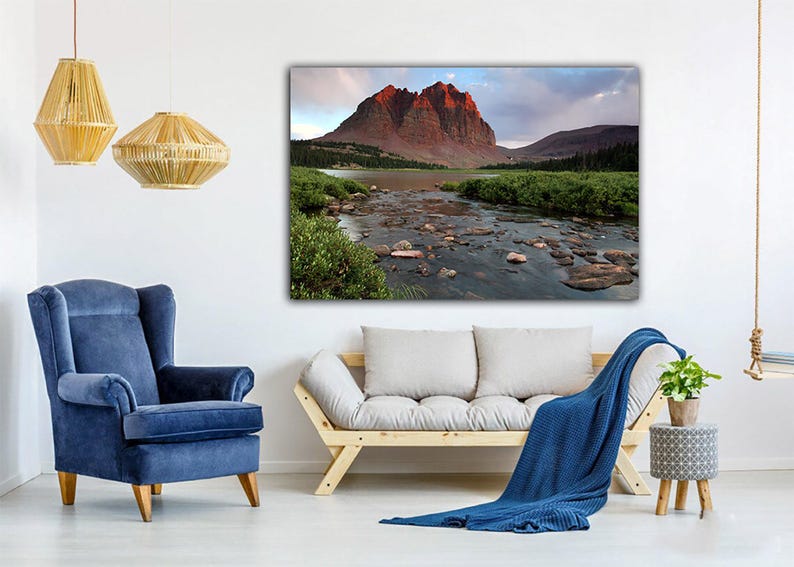 a living room with a large landscape painting of a mountain river and lush greenery hanging on the wall. The room is furnished with a blue armchair, a white sofa, and a wooden side table with a plant. The overall atmosphere of the room is calm and serene, with the natural scenery of the painting adding a touch of tranquility to the space.