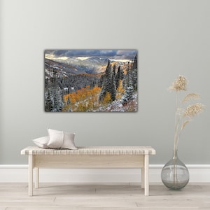 a large landscape painting hanging on a wall, depicting a mountainous, forested area with a mix of green, orange, and white colors. In front of the painting, there is a wooden bench with a single white pillow on it. To the right of the bench, there is a glass vase with dried flowers, adding a touch of natural beauty to the scene.
