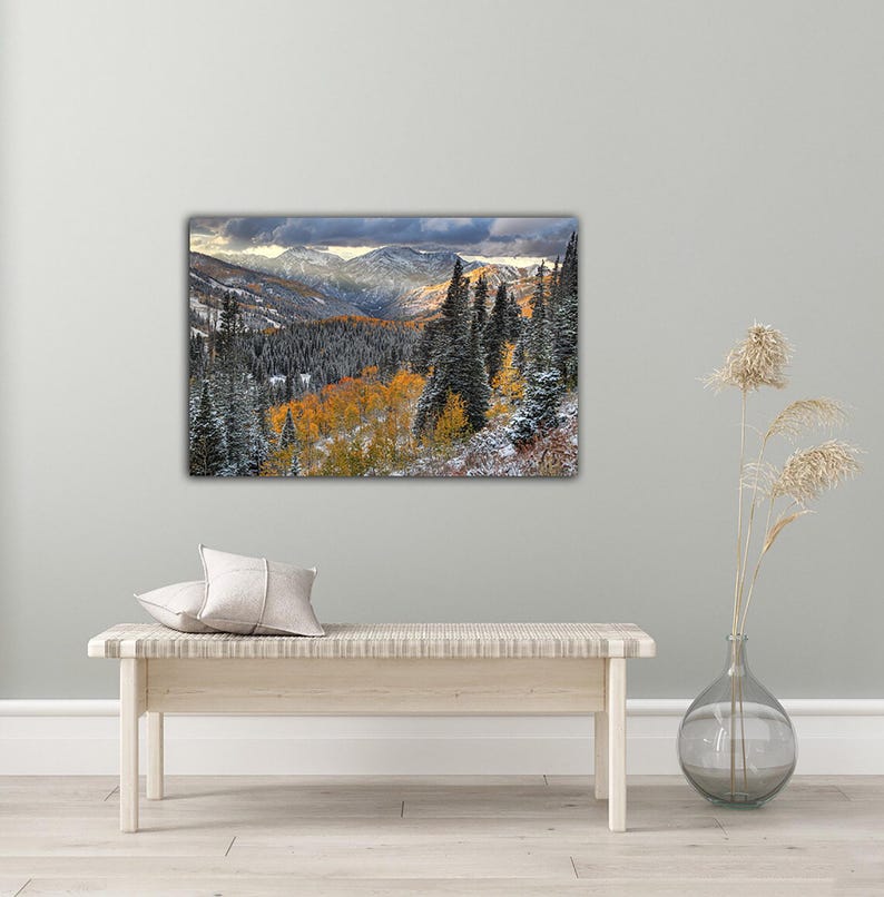 a large landscape painting hanging on a wall, depicting a mountainous, forested area with a mix of green, orange, and white colors. In front of the painting, there is a wooden bench with a single white pillow on it. To the right of the bench, there is a glass vase with dried flowers, adding a touch of natural beauty to the scene.