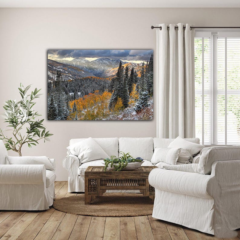 a serene living room with a large landscape painting hanging on the wall, featuring a snowy mountain range and colorful fall foliage. The room is furnished with a white sofa, a coffee table, and a potted plant, creating a cozy and inviting atmosphere.