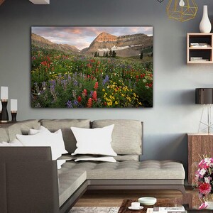 a large, vibrant painting of a mountain landscape with a colorful meadow in the foreground. The painting is hung on the wall above a gray sofa, creating a cozy and inviting atmosphere in the living room.
