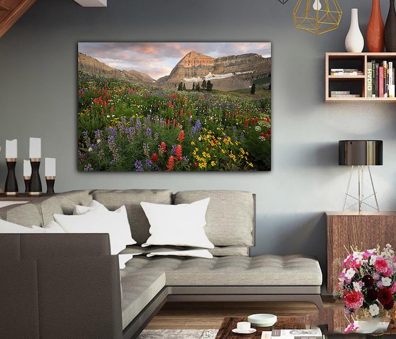 a large, vibrant painting of a mountain landscape with a colorful meadow in the foreground. The painting is hung on the wall above a gray sofa, creating a cozy and inviting atmosphere in the living room.