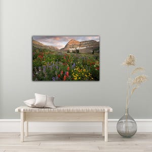 a large, vibrant painting of a mountain landscape with a colorful meadow in the foreground. The painting is hung on a wall above a wooden bench, which is positioned in front of a vase with dried flowers.