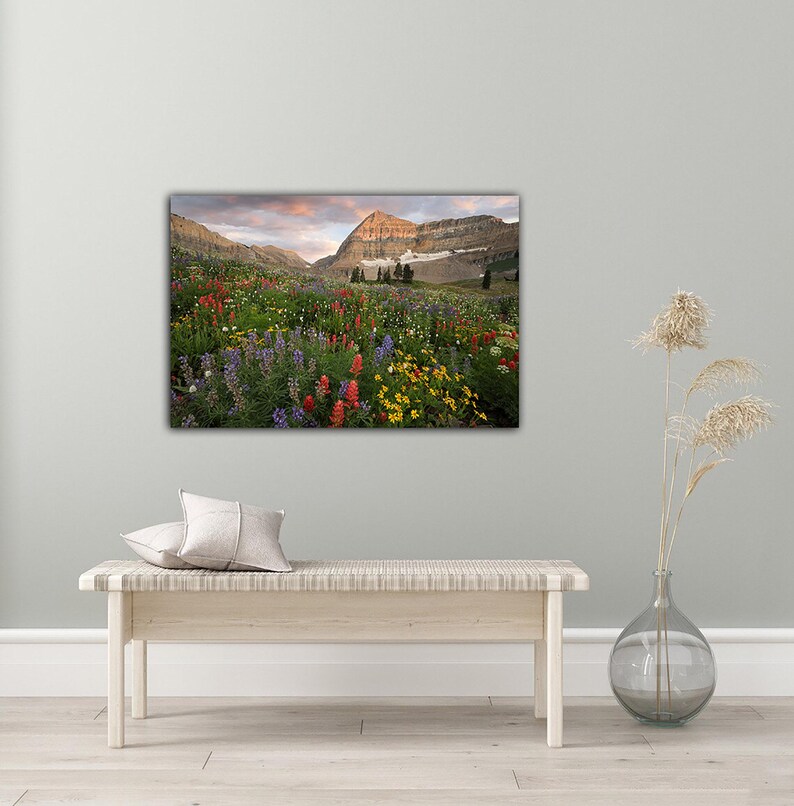 a large, vibrant painting of a mountain landscape with a colorful meadow in the foreground. The painting is hung on a wall above a wooden bench, which is positioned in front of a vase with dried flowers.