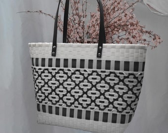 Puede incluir: Una bolsa de mano de tejido blanco y negro con asas negras. La bolsa está hecha de un material tejido y tiene un diseño geométrico.