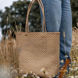May include: A woven straw tote bag with a light brown color. The bag has a wide, rectangular shape and is held by a person wearing blue jeans.
