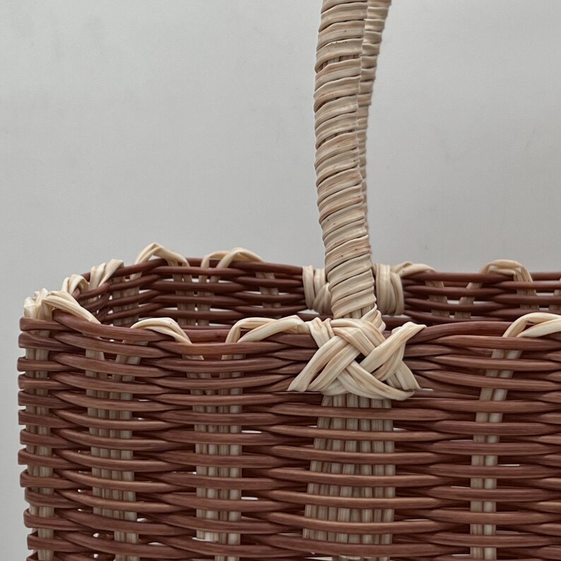 Puede incluir: Una cesta de mimbre tejida marr&oacute;n y crema con un solo asa. La cesta est&aacute; hecha de materiales naturales y tiene un aspecto r&uacute;stico.