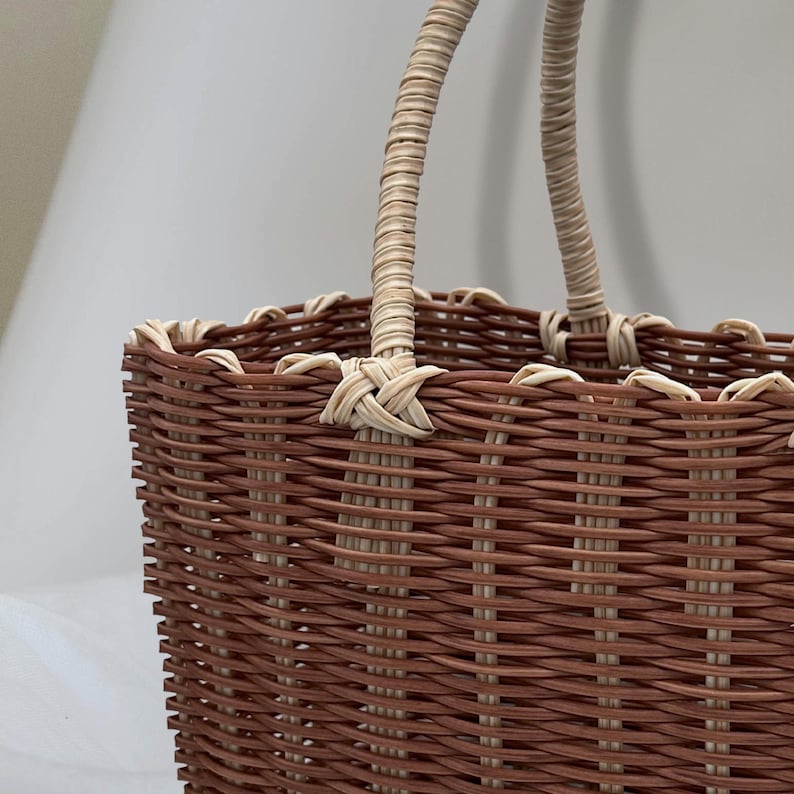 Puede incluir: Una cesta tejida de color marr&oacute;n y crema con dos asas de color crema. La cesta est&aacute; hecha de un material tejido y tiene una forma rectangular.