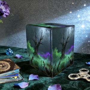 May include: A cube-shaped storage box with a spooky forest scene featuring bare trees, zombie hands, and green and purple mist. The box is on a green surface with playing cards, a die, and a fidget spinner.