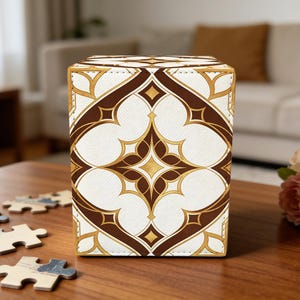 May include: A decorative box with a geometric pattern in brown, gold, and white. The box is rectangular and sits on a wooden table. Puzzle pieces are scattered near the box, and a bouquet of flowers is in the background.