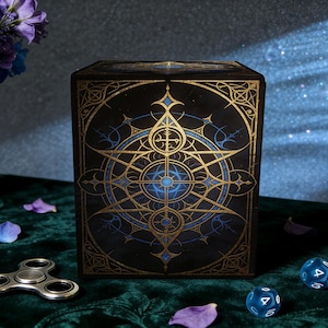 May include: A black box with a gold and blue geometric design. The box is decorated with intricate lines and shapes. A silver fidget spinner, blue dice, and flowers are on a green surface.