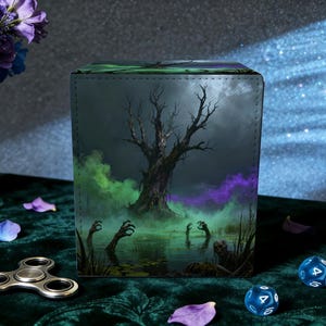 May include: A square, dark-themed dice box featuring a swamp scene with a bare tree, green and purple mist, and zombie hands reaching out of the water. The box is on a green surface with dice and a fidget spinner.