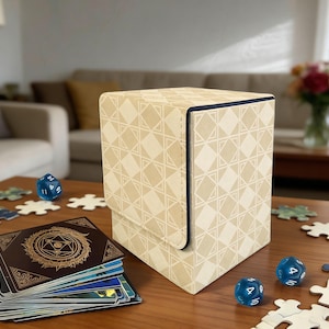 May include: A beige card storage box with a geometric pattern, blue dice, and a stack of cards with a gold design are on a wooden table. Puzzle pieces are scattered around the box and dice. The box has a dark blue trim.