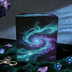May include: A black box with a swirling teal and purple galaxy design. The box is rectangular and stands upright on a dark green surface. A silver fidget spinner and two blue dice are in the foreground.