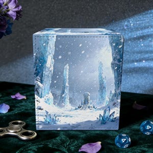 May include: A cube-shaped card storage box with a winter landscape design. The artwork depicts a snowy scene with icy formations and a distant structure. The box is on a green surface with dice, a fidget spinner, and flower petals.