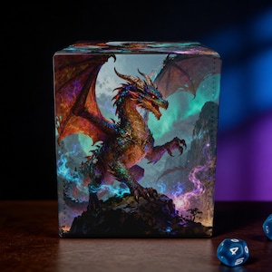 May include: A cube-shaped storage box featuring a detailed illustration of a dragon with red, orange, and blue scales, perched on a rocky landscape. The dragon has large wings and is set against a backdrop of a night sky. Two blue dice with white numbers are in the foreground.