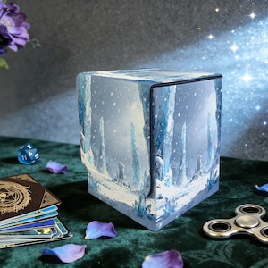 May include: A cube-shaped card storage box with a winter landscape design, featuring snow-covered mountains and a castle. The box is open, revealing the interior. A deck of cards, a die, and a fidget spinner are also visible.