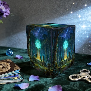 May include: A cube-shaped decorative box with a fantasy forest scene. The box features a glowing structure in a forest setting under a starry night sky. A deck of cards, a die, a fidget spinner, and flower petals are also visible.