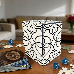 May include: A white and black patterned game card storage box with a magnetic closure sits on a wooden table. Blue dice, game cards, and puzzle pieces are scattered around the box. The box has a black trim.
