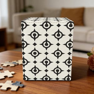 May include: A square, cream-colored box with a repeating black geometric pattern. The pattern consists of circles and diamond shapes. The box is sitting on a wooden table, with puzzle pieces scattered around it.