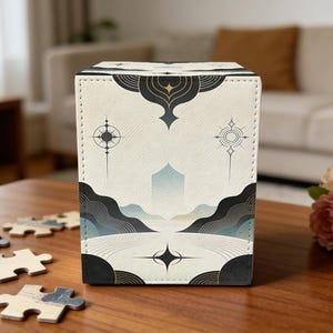 May include: A cream-colored box with a geometric landscape design featuring mountains, a central structure, and star-like elements. The box has black accents and is sitting on a wooden table with puzzle pieces.