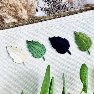 May include: A close-up of a white canvas pocket with four embroidered leaf patches in white, green, black, and light green. The pocket is part of a larger tan fabric item. Green leaves are in the foreground.
