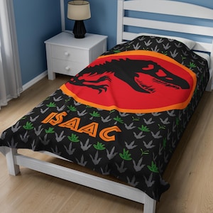 May include: Black twin-size bedspread with a dinosaur footprint pattern. The center of the bedspread features a red circle with an orange outline and a black silhouette of a dinosaur. The name "ISAAC" is printed in green and orange letters on the bedspread.