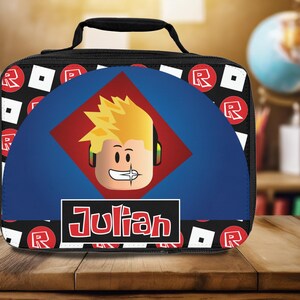 Pu&ograve; includere: Sacca da pranzo nera con un design blu e rosso che presenta un personaggio dei cartoni animati con i capelli biondi e il nome "Julian" stampato in rosso.