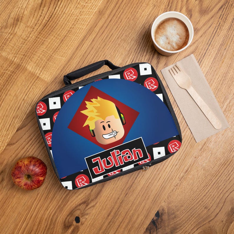 Pu&ograve; includere: Una borsa pranzo blu con un motivo a scacchi bianco e nero e loghi "R" rossi. La borsa presenta una grafica di un personaggio di Roblox con capelli biondi e cuffie, e il nome "Julian" in lettere rosse.