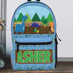 May include: A light blue backpack featuring a dinosaur theme. The top panel displays a landscape with green trees and three cartoon dinosaurs. The lower panel has the name "ASHER" in green block letters on a brown background. A black zipper and mesh side pocket are visible.